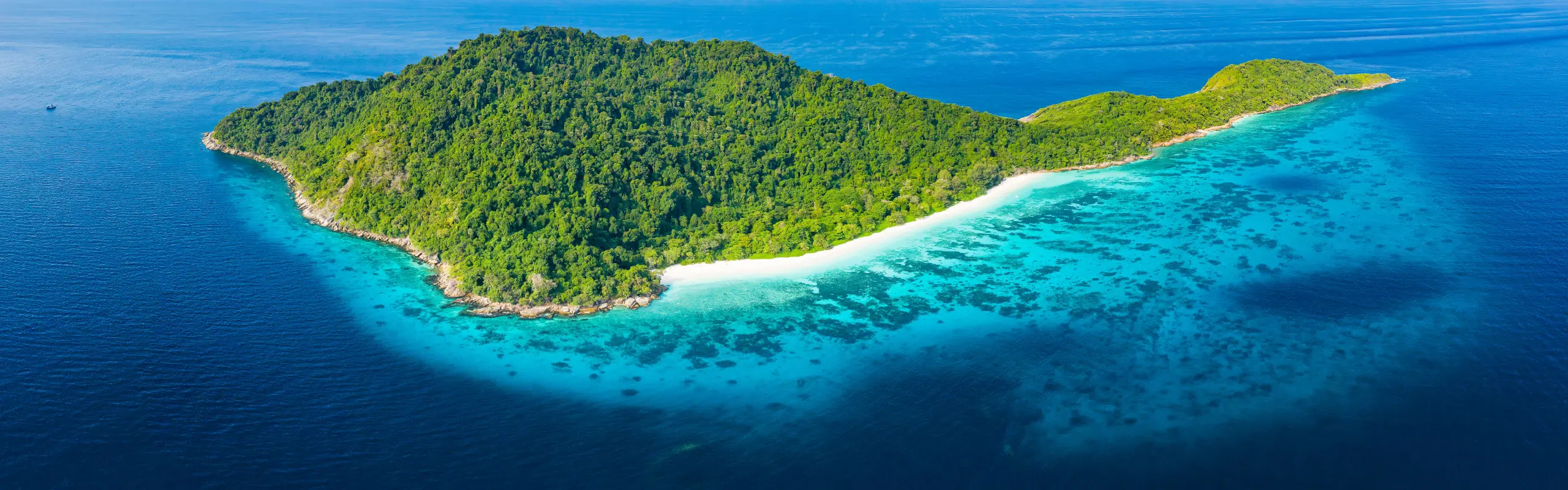 Aerial view of a forested island with a white sand beach and turquoise coral reef in the Andaman Sea